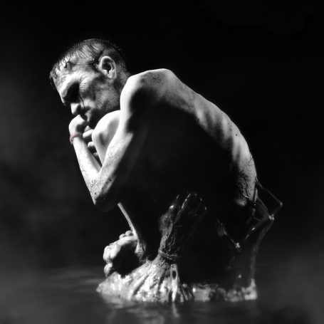 imagen en blanco y negro de modelado 3d de hombre estilo Frankestein, muy flaco, triste, desnudo y acurrucado, rodeado de agua y manos 